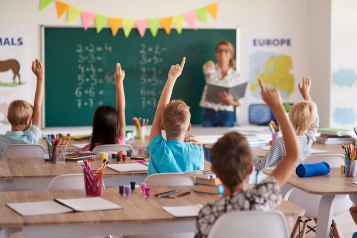 Nell'immagine alcuni alunni ripresi di spalle alzano la mano per rispondere alle domande della maestra - Smart Marketing