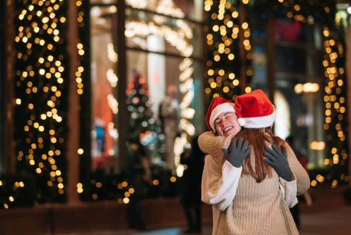 Nell'immagine due ragazze con li cappello di Babbo Natale si abbracciano per la strada di uan città addobbata a festa - Smart Marketing